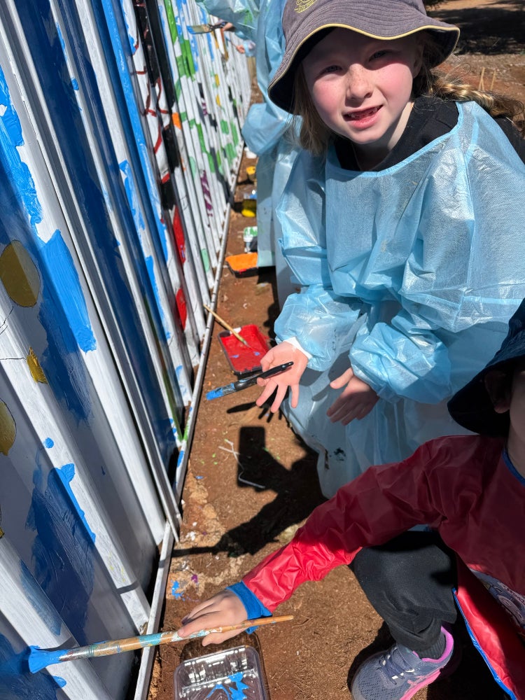 photo of a young girl holding a paint bush, helping to paint a mural on a storage container.